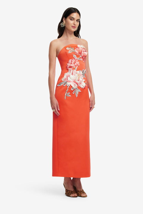 Woman posing in  an orange strapless maxi length dress with floral patterns on a white background