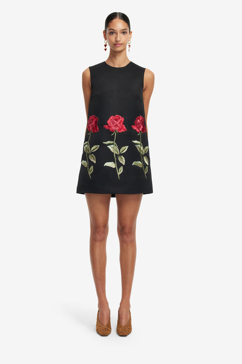 Woman wears a black sleeveless mini 
dress with red floral embroidery on a white background