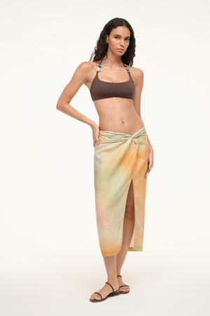 Woman wearing a pastel multi color midi skirt that features a twisted waistband detail and a deep front slit, worn over a bikini.