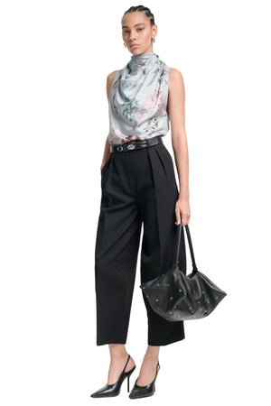 Woman wearing a bias cut sleeveless floral top and black pants, holding a black handbag on a white background.
