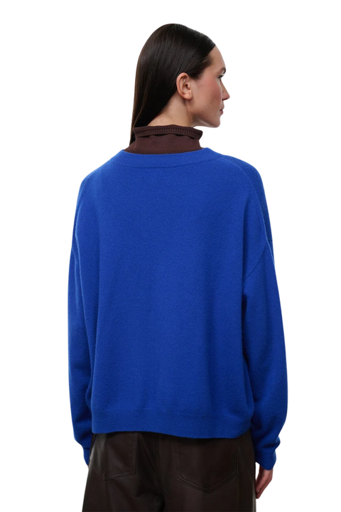 A woman shown from the back wearing a vibrant royal blue knit sweater over a dark brown mock-neck top, paired with dark brown trousers, against a solid white background.