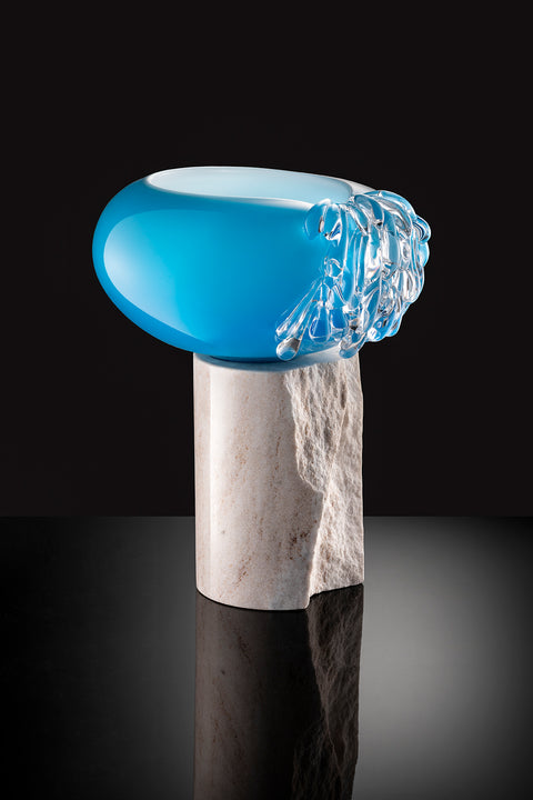 Different view of blue glass art piece atop light colored marble stand.