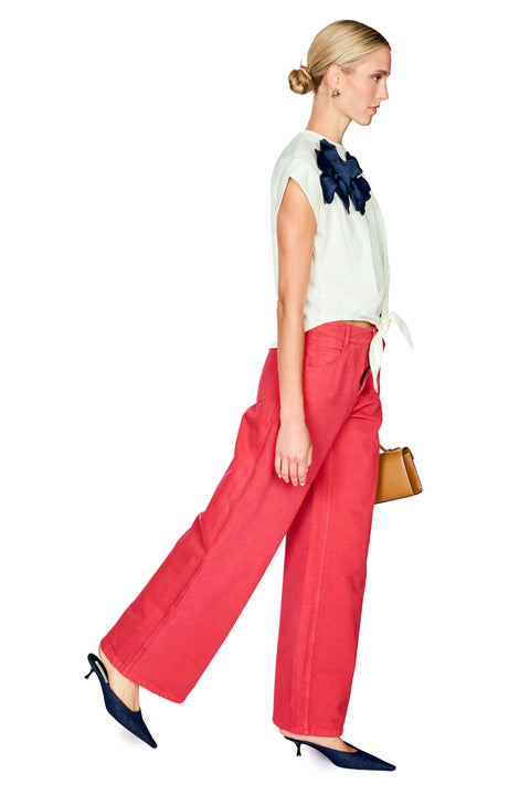 Woman wearing a white top with a navy floral detail and red pants on a white background side angle