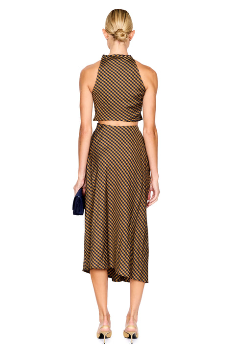 A model stands against a white background wearing a brown and navy checkered halter crop top and matching draped midi skirt, paired with beige pointed-toe heels and holding a black clutch.