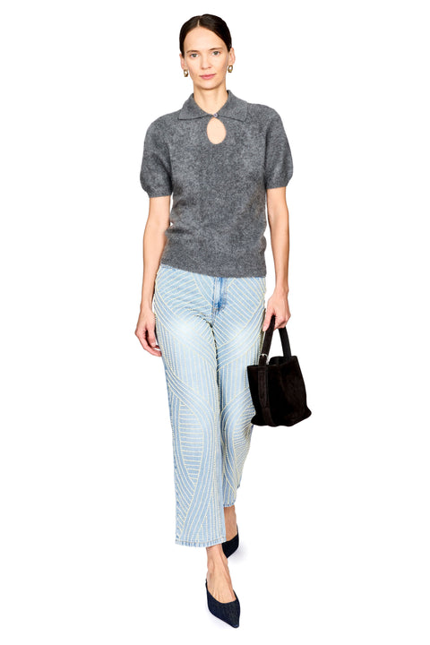 A model stands against a white background wearing a dark gray short-sleeve knit top with a keyhole cutout, light blue jeans with pearl detail, dark navy pointed shoes, and holding a dark brown handbag.