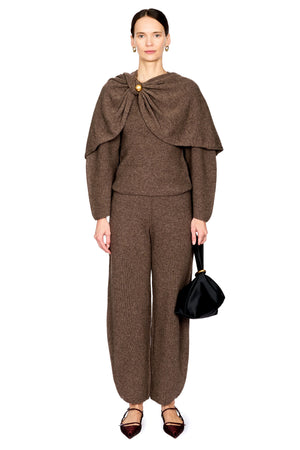 A model wearing a brown knit matching set consisting of a long-sleeve top with a draped collar and broch, and matching trousers, styled with dark pointed flats and a black handbag against a white background.