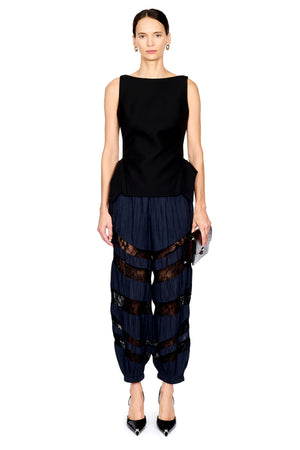 A model wearing a sleeveless black top and dark navy pleated harem-style pants with sheer black lace bands, standing against a plain white background.