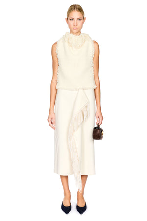 A model stands against a white studio background wearing a cream-colored sleeveless fringed top and matching fringed midi skirt, paired with dark navy pointed-toe heels and holding a small dark brown handbag.
