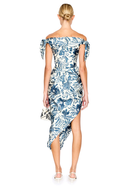 A woman viewed from the back wearing an off-the-shoulder midi dress with a blue and white tropical floral print. She is holding a matching clutch and wearing white high-heeled sandals against a plain white background.
