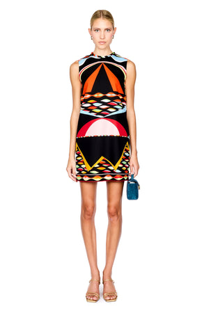 A model stands against a white background wearing a sleeveless black dress with a vibrant geometric and abstract print in orange, red, yellow, and light blue. She is holding a small blue handbag and wearing gold high-heeled sandals.
