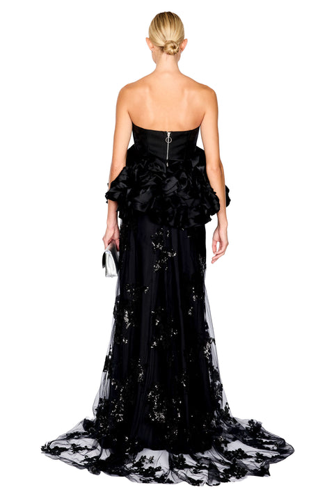 A model wearing a strapless top with a dramatic ruffled bodice, a long black skirt with a sheer floral sequin embellished lace overlay, posed against a plain white studio background.