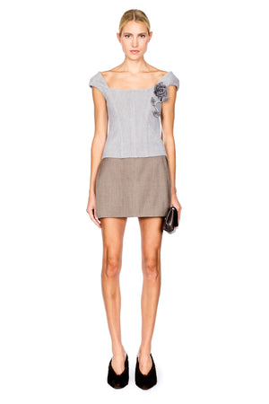A model stands against a white background wearing a light gray off-the-shoulder top with a dark floral embroidery on the sleeve and a taupe mini skirt, paired with black pointed shoes and holding a small black clutch.