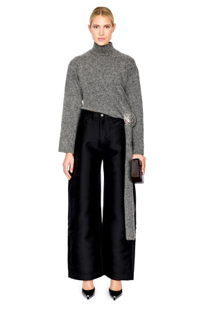A model stands against a white background wearing a gray cropped turtleneck sweater with a long tie detail, black wide-leg trousers, and black pointed shoes, while holding a small black clutch.