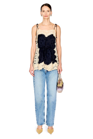 A model stands against a white background wearing a sleeveless top with a dark navy velvet ruffle and beige lace trim, paired with light-wash blue jeans and beige heels, holding a small patterned handbag.