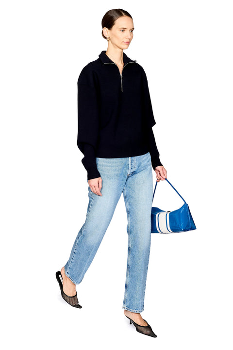 A woman modeling a navy long-sleeve quarter-zip sweater, light-wash blue jeans, and black pointed shoes, while holding a shiny blue handbag with white stripes against a plain white background.