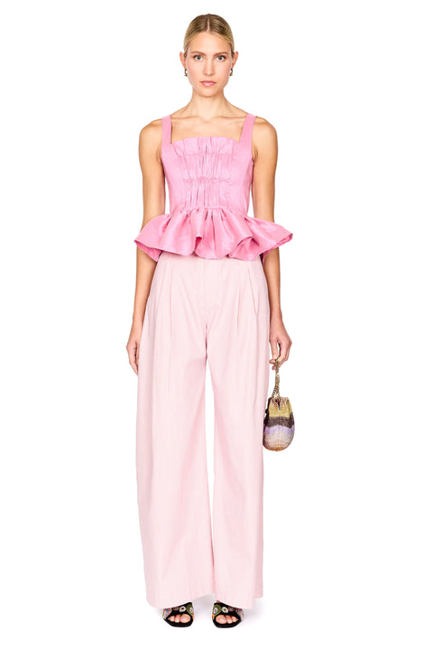 A model stands against a white background wearing a light pink two-piece outfit consisting of a smocked sleeveless top with a ruffled hem and matching wide-leg trousers. She is holding a small handbag.