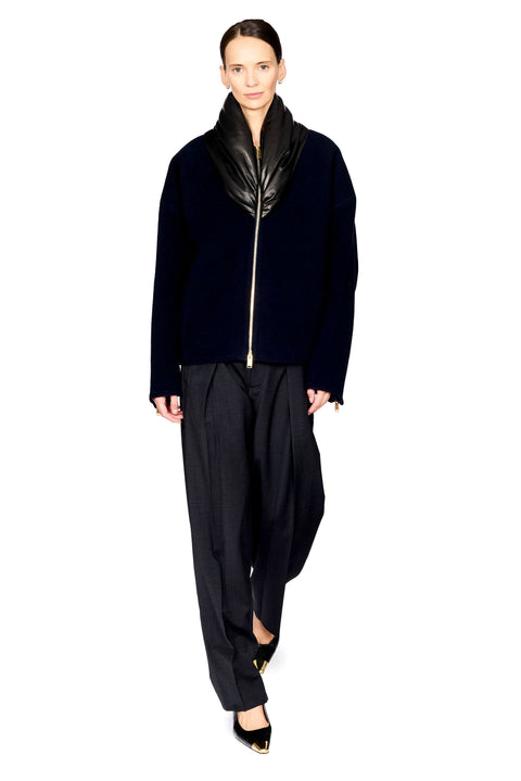 A model wearing a black cropped jacket with a high glossy black collar and a silver front zipper, paired with loose black trousers and black pointed shoes with gold accents, on a solid white background.