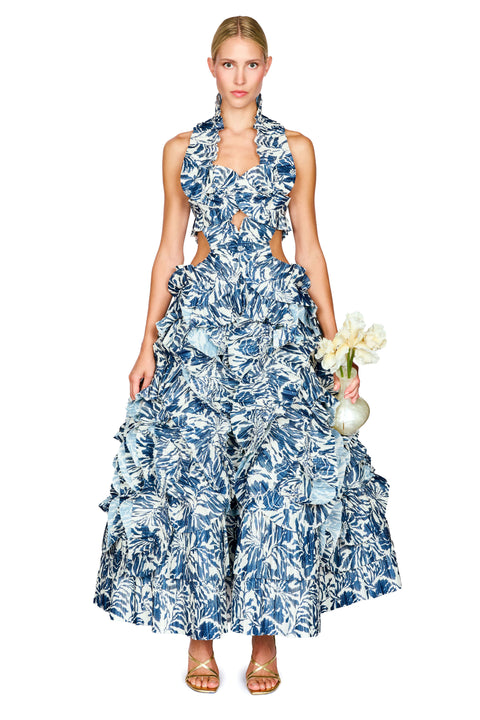 A model wearing a sleeveless halter-neck maxi dress with a blue and white tropical floral print, featuring side cutouts and a tiered ruffled skirt, holding a small vase of white flowers against a solid white background.