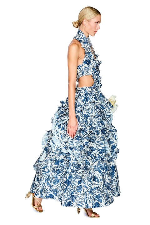A model wearing a blue and white tropical leaf print halter-neck ruffled maxi dress with a cutout waist, shown from a side profile against a white background.