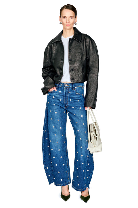 A model stands against a white background wearing a black leather jacket, blue wide-leg jeans with white polka dots, a white top, and dark green pointed shoes, while holding a white tote bag.