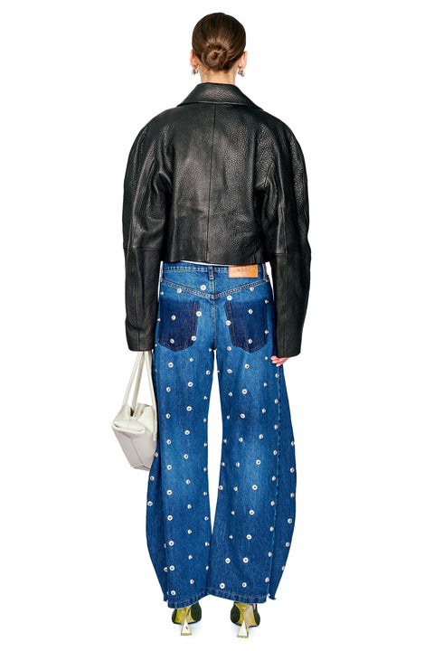 A model shown from the back wearing a black leather cropped jacket, wide-leg blue denim jeans with white floral embroidery, and holding a white shoulder bag against a plain white background.