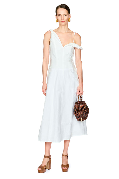 A model wearing a white one-shoulder midi dress with a ruffled off-the-shoulder detail, holding a brown woven handbag and wearing brown platform sandals against a plain white background.