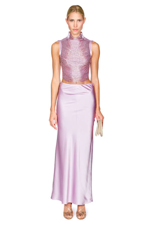 A model wearing a two-piece lavender formal set consisting of a sleeveless high-neck textured top and a matching satin skirt, paired with metallic heels and holding a small clutch, on a white background.