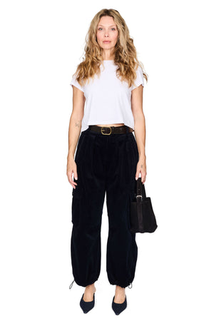 A woman modeling a white short-sleeve t-shirt, black velvet wide-leg cargo pants with a brown belt, holding a black handbag against a plain white background.