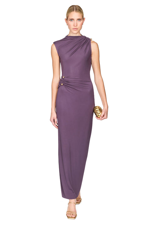 A woman modeling a sleeveless, floor-length draped gown in a muted plum-purple color, paired with gold sandals and holding a small gold clutch, set against a plain white background.