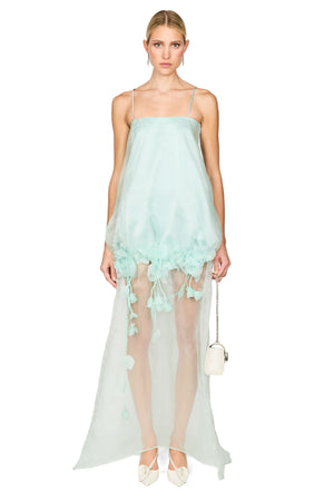 A model wearing a light mint green sheer maxi dress with spaghetti straps, a voluminous gathered skirt with tassel details, and holding a small white handbag against a solid white background.