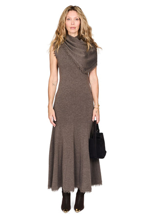 A woman modeling a sleeveless, high-neck, floor-length brown dress with a draped shawl over one shoulder and a frayed hem, paired with dark ankle boots and holding a black handbag against a plain white background.