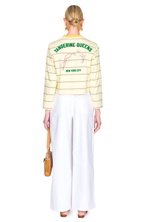 Woman wearing a yellow striped sweater with "tangerine queens new york city" on the back with white pants on a white background