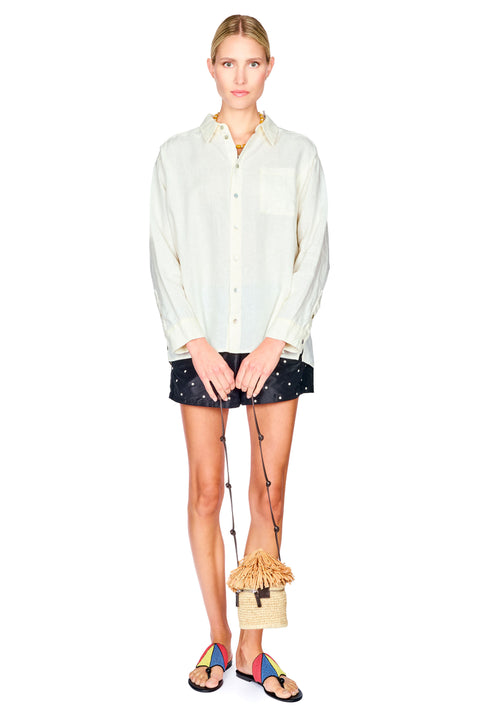 A model stands against a white background wearing a cream-colored long-sleeve button-down shirt, black shorts with white polka dots, and colorful sandals, while holding a small woven straw handbag with a long strap.