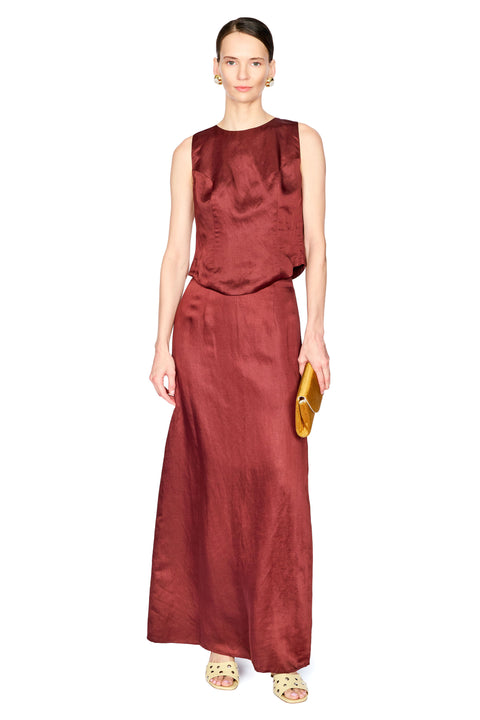 A model wearing a sleeveless, floor-length rust red two piece set, holding a small gold clutch against a plain white background.