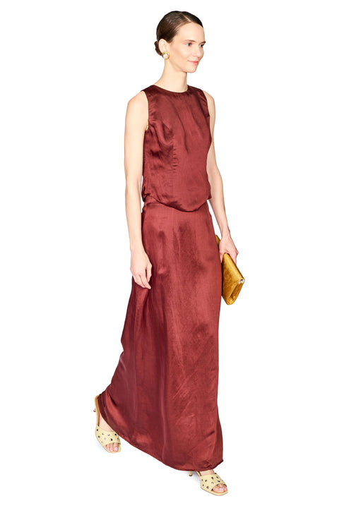 A woman modeling a sleeveless, high-neck, two piece set in a deep burgundy red. She is holding a small gold clutch and wearing gold sandals, set against a plain white background.