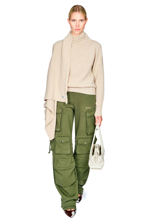 A model wearing a beige knit cowl-neck sweater with a draped scarf, olive green cargo pants, and carrying a white tote bag, set against a plain white background.