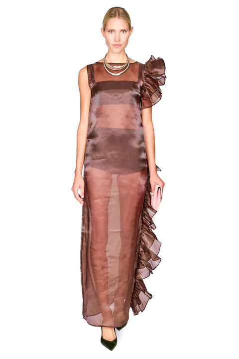 A model wearing a sheer brown maxi dress with a ruffled one-shoulder design, layered necklaces, and dark green pointed shoes, set against a plain white background.