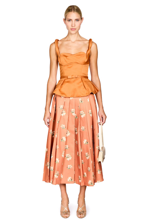 A model stands against a white studio background wearing a two-piece outfit consisting of a cropped, ruffled orange-brown top and a pleated peach skirt with a white floral pattern. She is holding a small beige handbag and wearing strappy high heels.