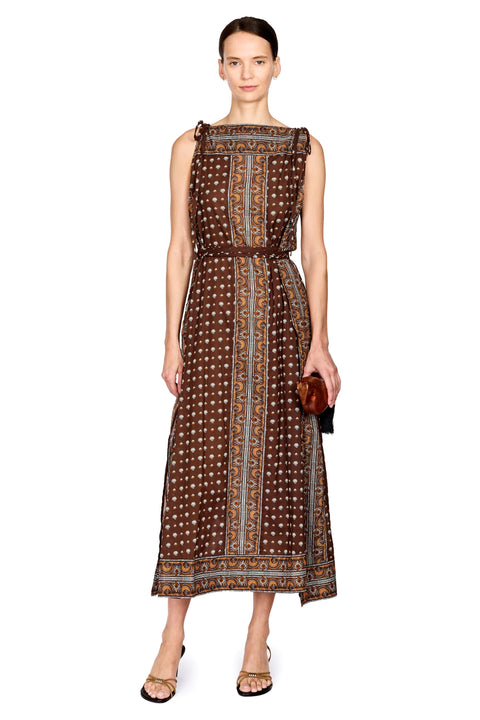 A woman modeling a sleeveless midi dress with a brown base, white polka dots, and vertical orange and blue patterned bands, styled with a matching belt and holding a small brown clutch against a plain white background.