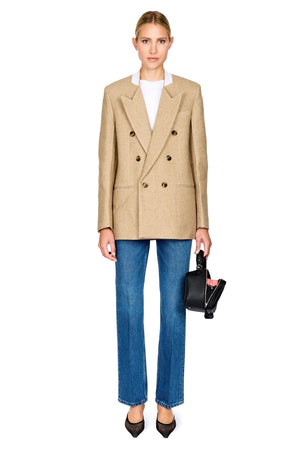 A model stands against a white background wearing a beige double-breasted blazer over a white top, blue jeans, and black heels, while holding a small black handbag.