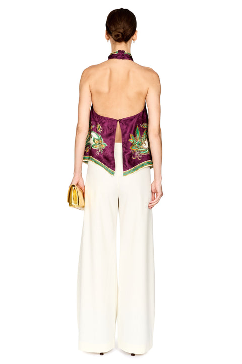A model shown from the back wearing a halter-style top with a deep open back, featuring a deep purple silk-like fabric with green and gold embroidery, paired with wide-leg cream trousers and holding a small gold clutch.