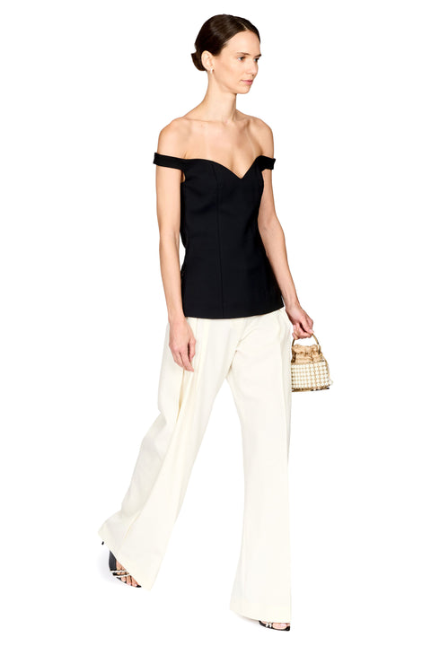 A woman modeling an off-the-shoulder black top and off-white wide-leg pants, holding a small woven handbag against a plain white background.