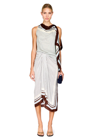 A model wearing a sleeveless draped midi two piece set with a white and brown geometric pattern, featuring a wrap-style skirt and a matching draped scarf, holding a small navy blue clutch against a white background.