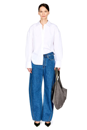 A model stands against a white studio background wearing an oversized white button-up shirt, wide-leg blue jeans, black pointed shoes, and holding a large gray tote bag.