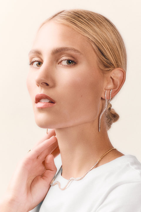 A close-up portrait of a woman wearing elegant wavy jewelry, featuring a delicate gold necklace and matching wavy drop earrings with sparkling clear stones, set against a soft beige background.