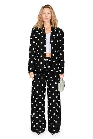 A woman stands against a plain white studio background wearing a black wide-leg blazer and matching trousers with large white polka dots, paired with a white cropped top and holding a small light gray handbag.