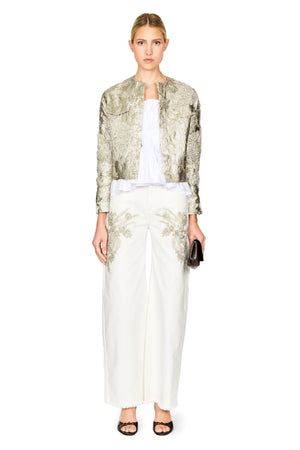 A model stands against a white studio background wearing a metallic gold and silver patterned cropped jacket over a white top, paired with wide-leg white trousers and black sandals.