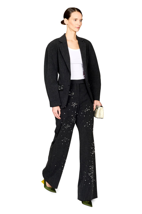 A model wearing a black tailored blazer and matching wide-leg trousers with scattered silver embellishments, styled with a white top, olive green heels, and holding a small metallic clutch against a plain white background.