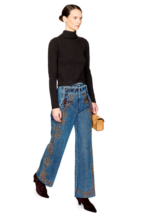 A model wearing a black turtleneck sweater, blue wide-leg jeans with ornate gold embroidery down the sides, and dark brown pointed-toe heels, holding a small tan handbag against a plain white background.