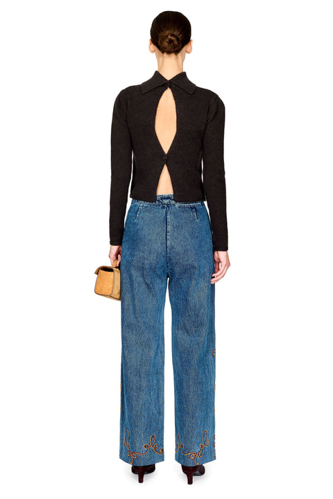 A model shown from the back wearing a black long-sleeve knit top with a dramatic open back and wide-leg blue jeans with gold embroidery near the hems, holding a small tan handbag against a white background.